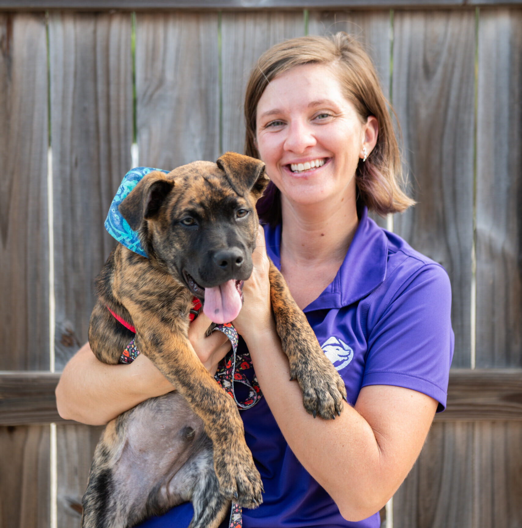 Smiling dog trainer from Happy With Dogs holding a brindle puppy wearing a blue bandana and red harness, standing in front of a wooden fence.